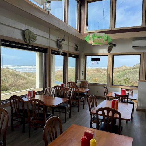 The image shows a cozy restaurant with wooden tables, large windows, and views of sand dunes and the ocean outside.