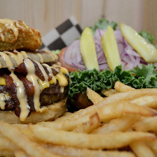 The image shows a cheeseburger topped with cheese sauce, bacon, and a fried item, accompanied by lettuce, pickles, onion slices, and a pile of fries.