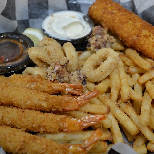 The image shows a basket containing fried shrimp, calamari, fries, and a fried fish fillet, served with dipping sauces on a checkered paper lining.