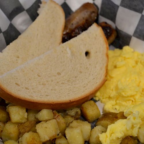 This image shows a breakfast plate with scrambled eggs, two slices of bread, diced potatoes, and sausages, served on a checkered paper liner.