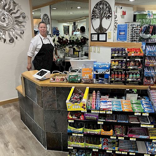 A person behind a counter with various snacks and items for sale is shown. There are also sunglasses and a decorative wall piece.