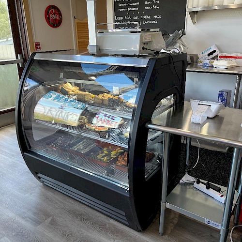 A display case filled with various food items is next to a countertop with a cash register in a small cafe or bakery setting.