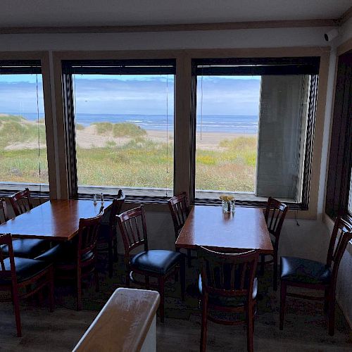 A cozy dining area with wooden tables and chairs overlooks a beach through large windows, offering a serene view of sand dunes and the ocean beyond.