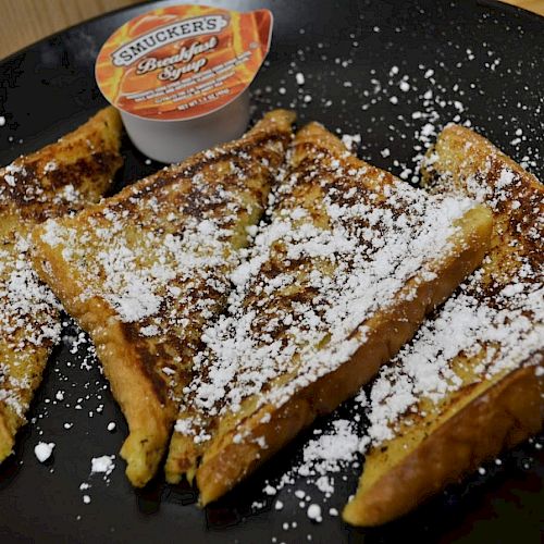 The image shows four slices of powdered sugar-dusted French toast on a black plate, accompanied by a small container of Smucker's syrup.