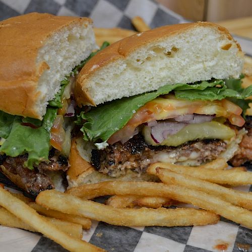 A cheeseburger sliced in half with lettuce, tomatoes, and onions, accompanied by a side of French fries on a checkered paper.