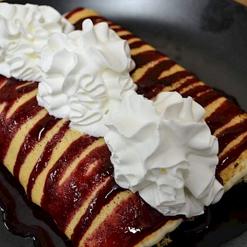 A crepe drizzled with berry sauce and topped with whipped cream is presented on a black plate.