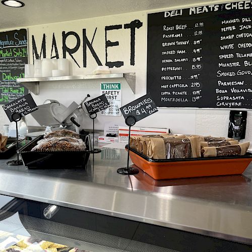 A deli counter with various breads and baked goods, chalkboard menus on the wall listing deli meats and cheese offerings.