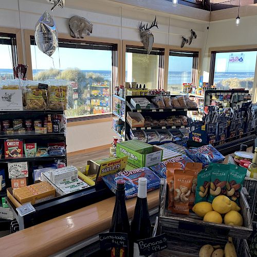 A small store by the beach features snacks, fresh produce, magazines, and hunting decor with large windows offering a scenic view of the ocean ending the sentence.