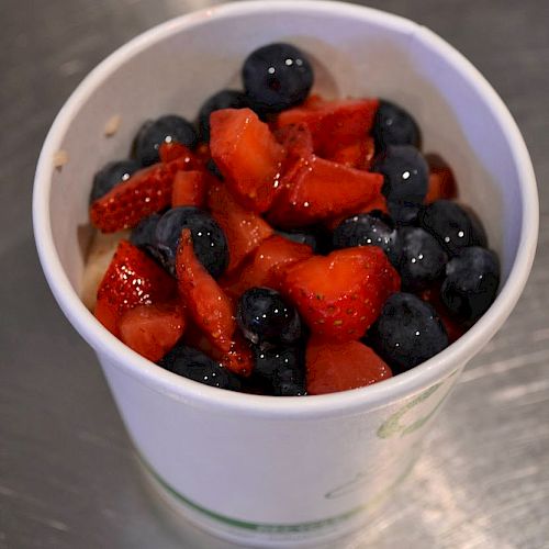 The image shows a white cup filled with sliced strawberries and blueberries on a metallic surface.