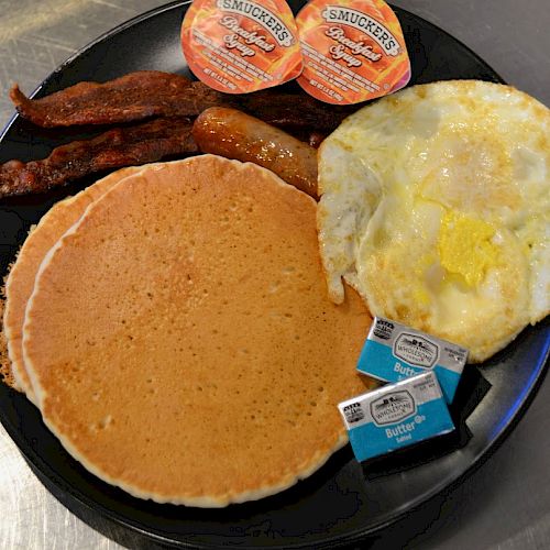 The image shows a breakfast plate with pancakes, syrup, bacon, a sausage link, fried eggs, and butter packets on a black plate.