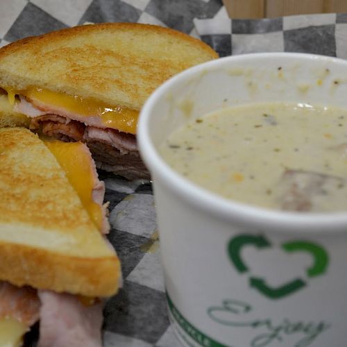 A grilled sandwich with ham and cheese is placed next to a cup of creamy soup on a checkered paper. Enjoy.