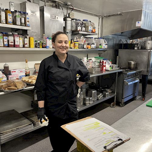 A person in a black chef uniform stands in a commercial kitchen with shelves of ingredients and cooking equipment around them, and a clipboard on a table.