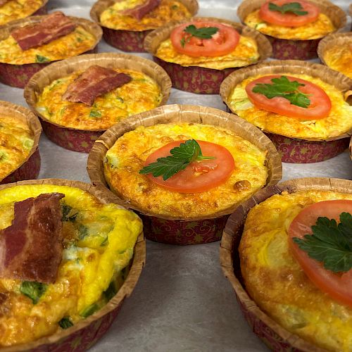 The image shows several mini quiches, some topped with slices of tomato and parsley, while others have pieces of bacon. They are placed on a baking tray.