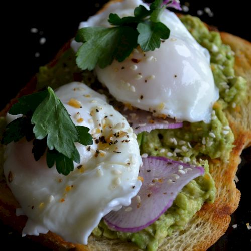 The image shows avocado toast topped with poached eggs, sliced onions, fresh parsley, and a sprinkle of black pepper on a slice of bread.