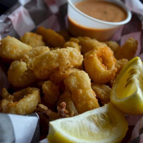 A basket of fried calamari with lemon slices and dipping sauce, served in a paper-lined basket.