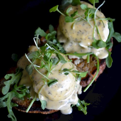 A dish featuring two poached eggs on toast, topped with creamy sauce and garnished with fresh greens, served on a dark plate.