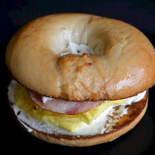 A bagel sandwich with layers of egg, ham, and cheese on a toasted bagel, viewed from the side against a dark background.