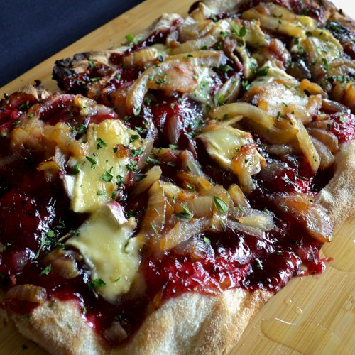 The image shows a flatbread topped with melted cheese, caramelized onions, herbs, and a red sauce on a wooden board.