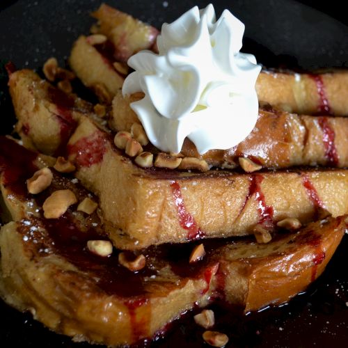 French toast sticks drizzled with syrup, topped with whipped cream and chopped nuts, on a dark plate.
