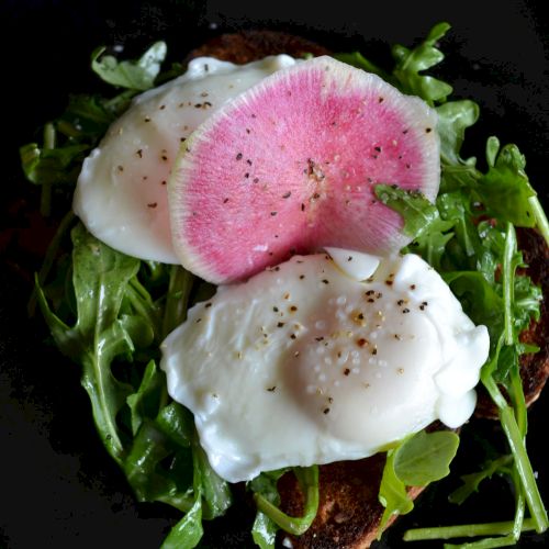 A dish featuring poached eggs, watermelon radish slices, and arugula on toast, garnished with black pepper.