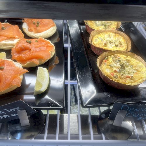 The image shows bagels with lox and Denver quiche displayed in a refrigerated case, each on a black rectangular plate, labeled with price tags.