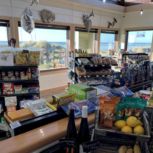 A small convenience store with snacks, drinks, fruit, and various items is shown. In the background, large windows offer a seaside view.