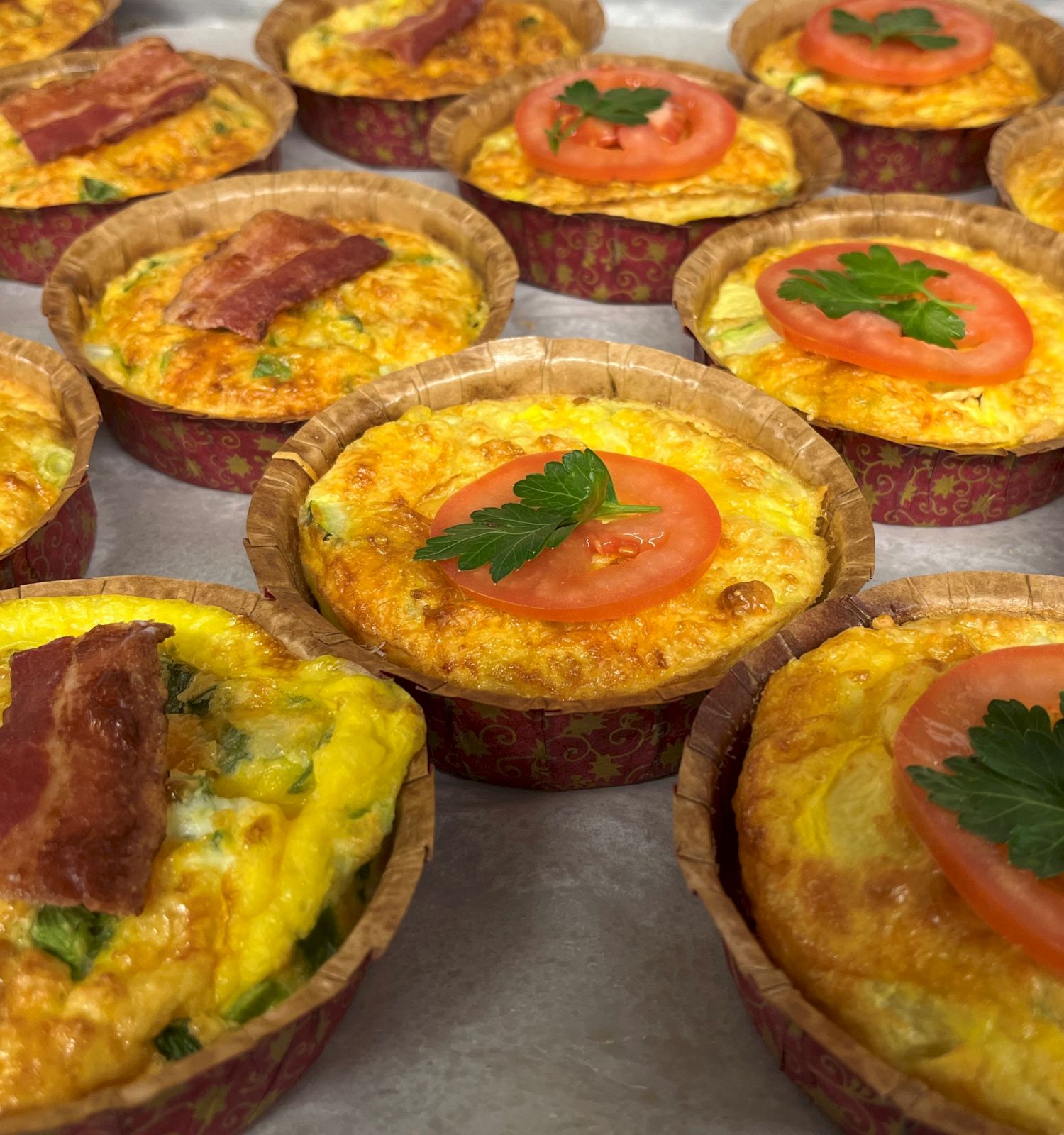 The image shows multiple baked quiches in cups, topped with either slices of bacon or tomato and a garnish of parsley, arranged on a table.