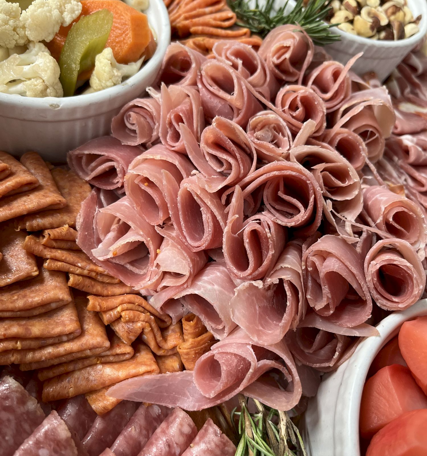 An assortment of charcuterie items, including sliced meats, crackers, pickled vegetables, and nuts arranged on a platter, with sprigs of herbs.