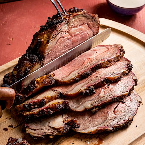 A person slicing a seasoned, roasted piece of meat on a wooden cutting board with a large knife and meat fork, showing several slices.