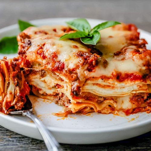 The image shows a slice of lasagna on a white plate with a fork, garnished with fresh basil leaves.