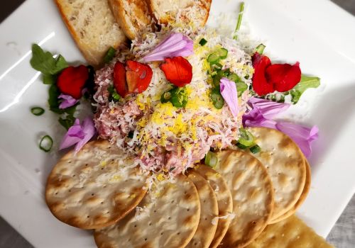 A plate with toasted bread, round and square crackers, cheese, shredded meat, strawberries, purple flowers, and greens in the center.