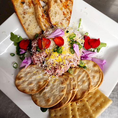 A plate with toasted bread, round and square crackers, cheese, shredded meat, strawberries, purple flowers, and greens in the center.