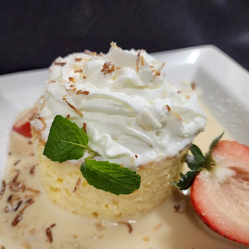 A dessert with whipped cream, a mint leaf, and a sliced strawberry on a white plate, drenched in sauce, sprinkled with toasted bits.