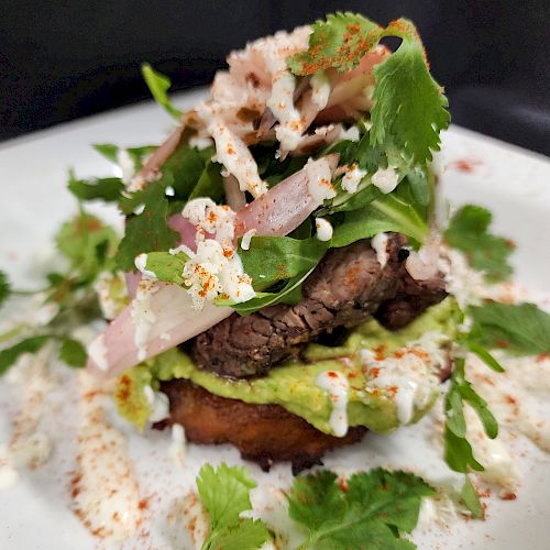 A gourmet dish with beef, guacamole, greens, and cheese on a fried base, garnished with cilantro and onions, served on a white plate.