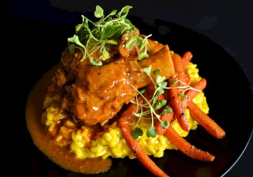 A dish featuring braised meat on a bed of saffron risotto with roasted carrots and microgreens, served on a black plate.