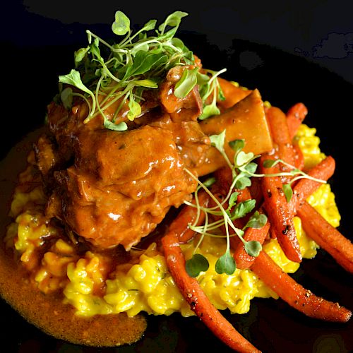 A dish featuring braised meat on a bed of saffron risotto with roasted carrots and microgreens, served on a black plate.
