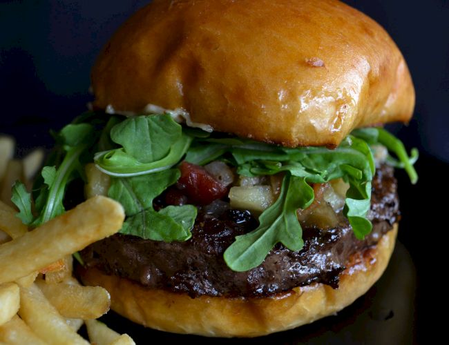 A juicy burger topped with greens, cheese, and bacon, beside crispy fries on a dark background.