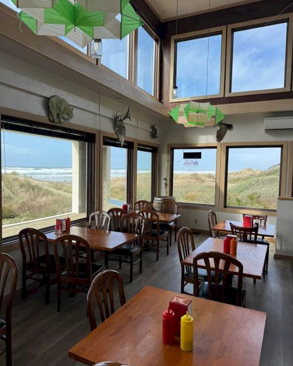 A seaside café with wooden tables and chairs, condiments on tables, large windows with ocean views, and green ceiling décor.